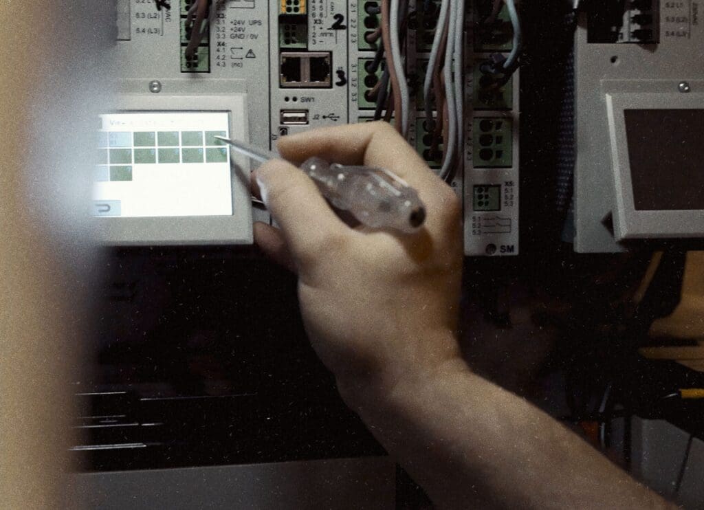 A person uses a screwdriver to interact with a touchscreen on an electrical panel, adjusting the natural ventilation system among various wiring and components.
