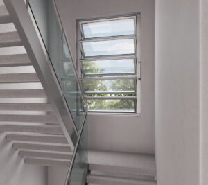 Stairwell with glass railings and modern horizontal windows showing an outdoor view. The walls are light gray, and the stairs are made of concrete.