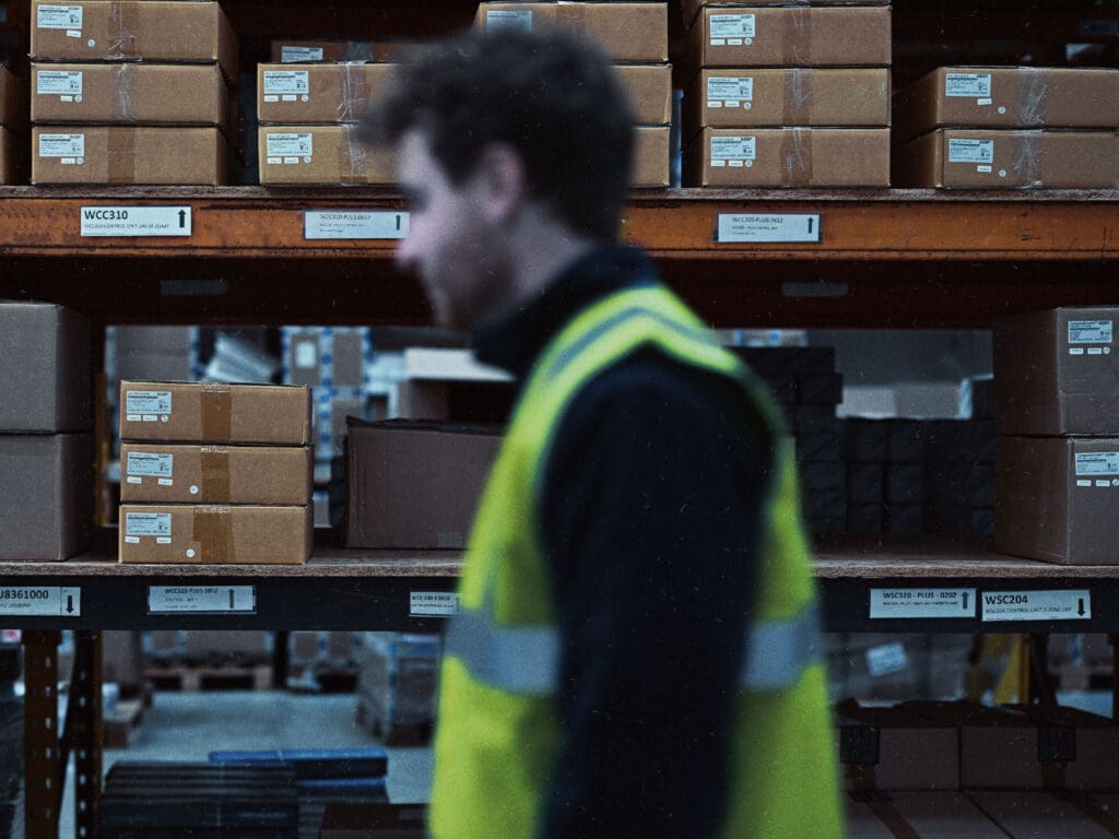 A worker in a reflective vest walks past shelves stacked with labeled cardboard boxes in a warehouse.