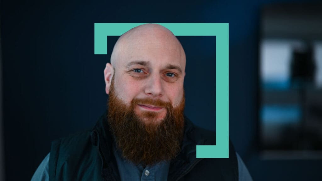 Portrait of a bald man with a full beard, smiling at the camera, framed by a turquoise geometric outline on a blurred indoor background.