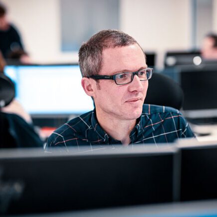 A man with glasses is seated at a desk in an office, concentrating on work. Other workers are seen in the background at their desks, enjoying the fresh air brought in by natural ventilation systems.