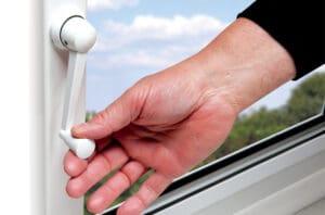 A hand is turning the white window handle to open or close a window with a sky and greenery visible outside, allowing for natural ventilation.