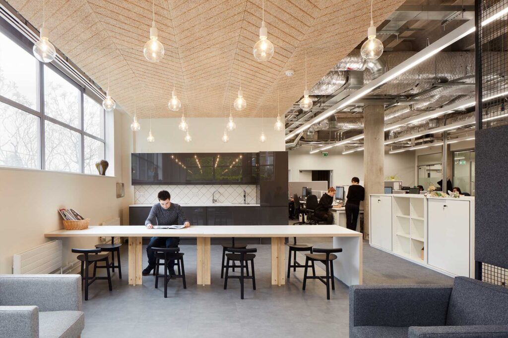 Modern office space with a high ceiling, hanging lights, and a long communal table. One person is working at the table while others are seen in the background at desks. Large windows provide natural light.