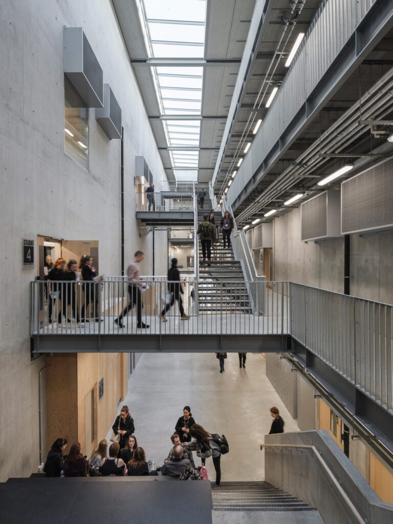 Interior view of a modern concrete building with multiple levels connected by stairways. Groups of people are walking, standing, and sitting on various levels. Skylights provide natural light.