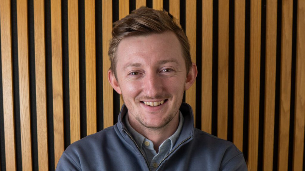 A person with short light brown hair, wearing a grey zip-up jacket and light shirt, smiles in front of a wall with vertical wooden slats.