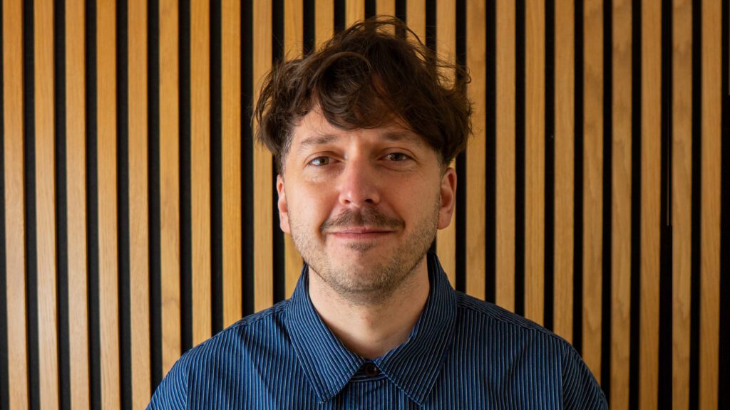 A man with wavy brown hair and light facial hair, wearing a blue collared shirt, stands in front of a wooden wall with vertical black stripes.