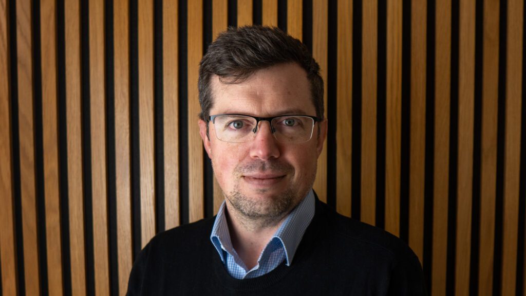 A man with glasses and short brown hair stands in front of a wooden slat wall, wearing a blue collared shirt and a black jumper.