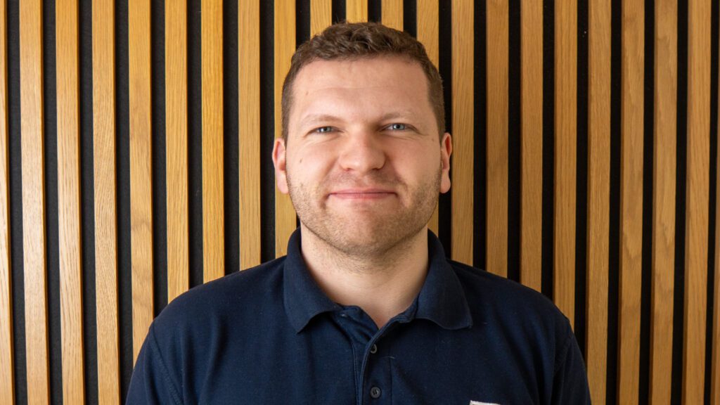 A man with short hair and a trimmed beard stands in front of a vertical wooden slat wall, wearing a dark polo shirt and smiling slightly at the camera.