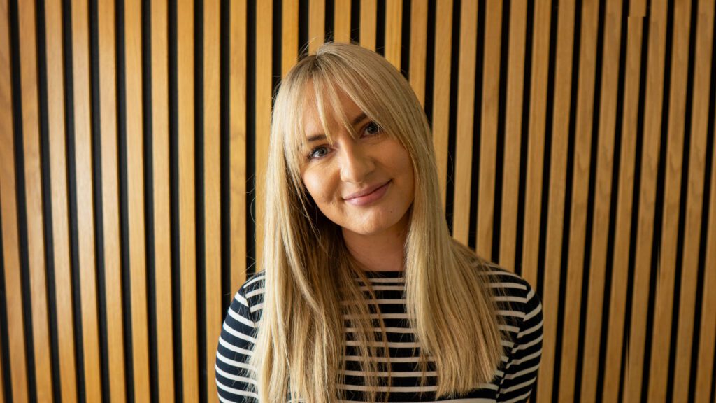 A person with long blonde hair, wearing a black and white striped shirt, stands in front of a wall with vertical wooden slats.