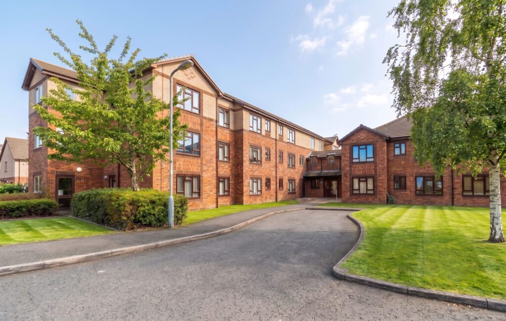 A three-storey brick block of flats with multiple windows, surrounded by trimmed lawns, trees, and a curved tarmac driveway under a clear sky.
