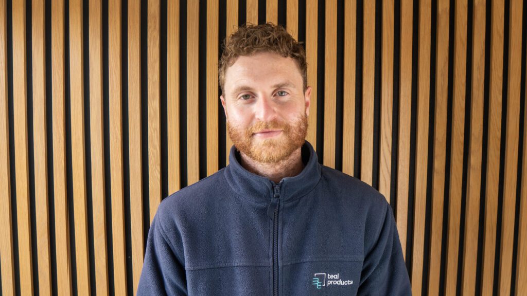 A man with curly hair and a beard stands in front of a wooden slat wall, wearing a navy fleece jacket with a “teal products” logo.