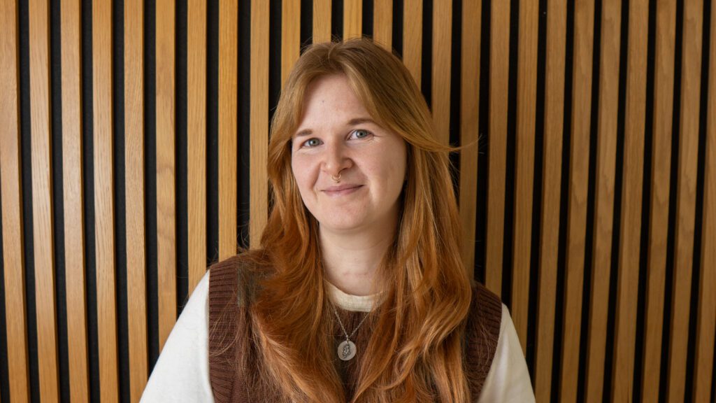 A person with long reddish hair and a brown waistcoat over a white shirt stands in front of a wall with vertical wooden slats, smiling slightly at the camera.