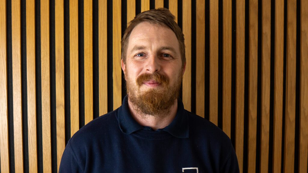 A man with a beard and moustache stands in front of a wall with vertical wooden slats, wearing a dark crewneck shirt.
