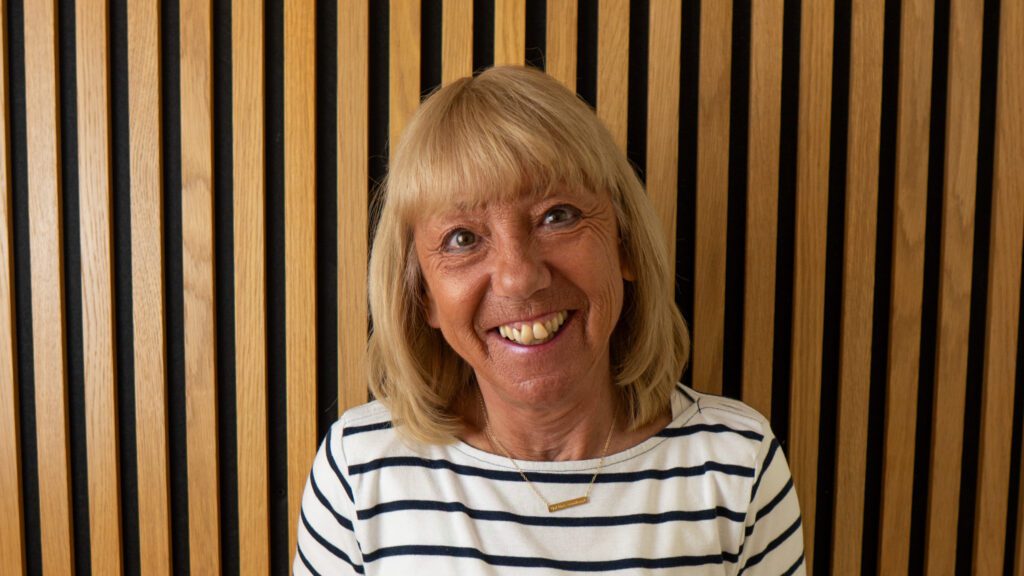 A woman with blonde hair and a striped shirt smiles in front of a wooden slat wall.