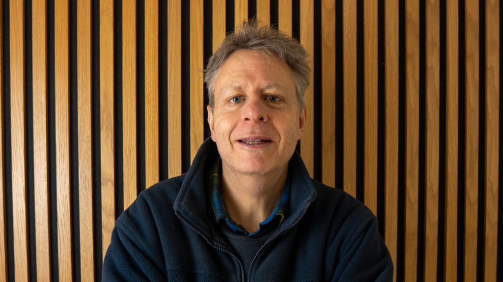 A middle-aged man with grey hair and a navy fleece jacket sits in front of a vertical wooden slat wall, facing the camera.